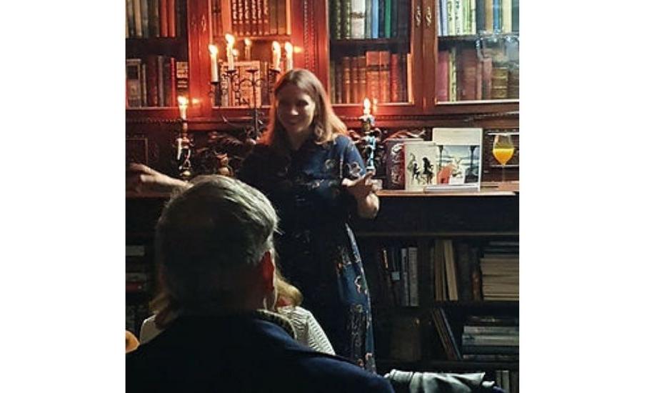 Dawn standing in front of a bookshelf in dim lighting smiling and telling stories to a crowd of people sitting around her.