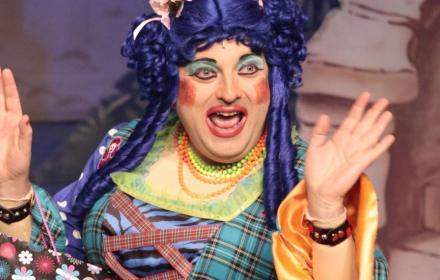 The picture shows a Pantomine Dame in heavy make up and blue wig, raising her hands at the audience